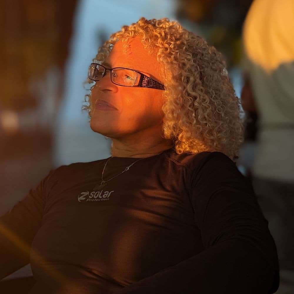 Jô em seu ateliê de costura, trabalhando com precisão em ajustes de roupas com ferramentas profissionais e tecidos ao fundo